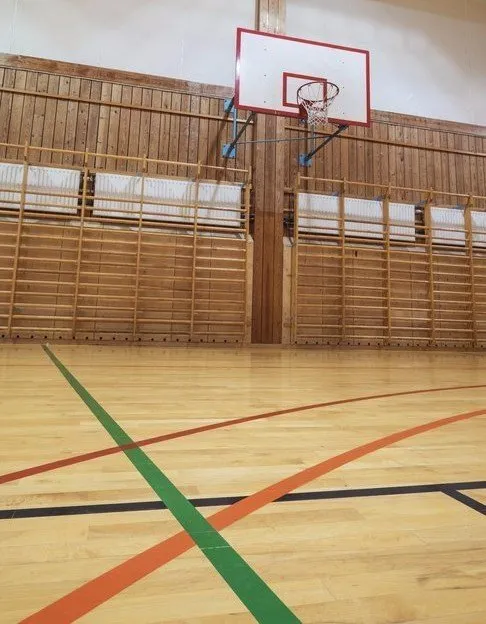 Indoor basketball court with a wooden floor, featuring a red and white basketball hoop and wall bars in the background.