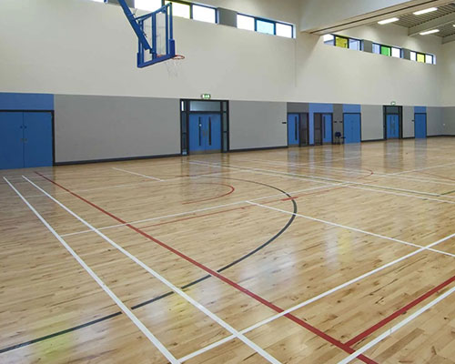 An empty indoor sports hall with a polished wooden floor, marked court lines, and a basketball hoop on the left.